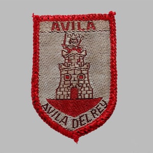 May include: A woven patch in a shield shape, featuring a red border and a central design. The design includes a castle tower and the text "AVILA" and "AVILA DEL REY" in red. The background is a light beige color.