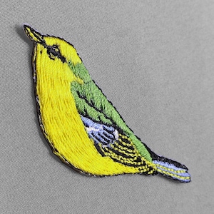 May include: Embroidered patch of a small bird with yellow, green, black, and blue thread. The bird is angled to the left, with its head and beak pointing upwards. The patch is on a gray background.