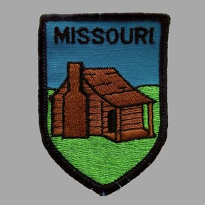 May include: Embroidered patch featuring the word "MISSOURI" above a brown cabin with a chimney, set against a blue sky and green grass. The patch has a black border and is shaped like a shield.