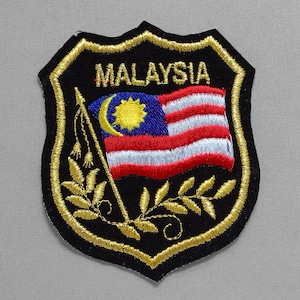 May include: Embroidered patch featuring the word "MALAYSIA" above a depiction of the Malaysian flag. The patch is shield-shaped with a black background, gold trim, and a gold leaf design.