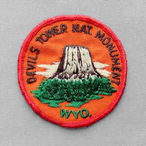 May include: A circular embroidered patch with an orange background and a red border. It features a depiction of Devils Tower with green trees at the base and white clouds. The text reads "DEVILS TOWER NAT MONUMENT" and "WYO." in dark brown.
