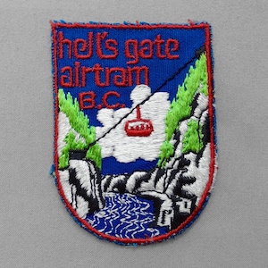 May include: Embroidered patch with a blue background and red border. The patch features the text "Hell's Gate Airtram B.C." in red, along with a depiction of a cable car, mountains, and a river. The patch has a textured appearance.