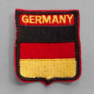 May include: Embroidered patch featuring the word "GERMANY" in gold thread above a black, red, and gold shield design. The patch has a red border and a shield shape, resembling the German flag.