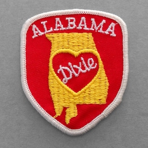 May include: A red and white embroidered patch with the outline of the state of Alabama in yellow. The word "Dixie" is embroidered in white inside a red heart in the center of the state outline. The words "Alabama" are embroidered in white above the state outline.