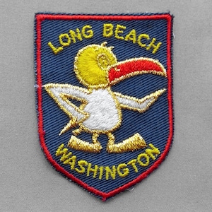 May include: A shield-shaped embroidered patch with a dark blue background and a red border. It features a yellow and white bird with a large red beak, surrounded by the yellow embroidered text LONG BEACH at the top and WASHINGTON at the bottom.