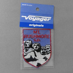 May include: A blue and red embroidered patch featuring the faces of the presidents from Mount Rushmore, with the text "MT. RUSHMORE S.D." The patch is in a clear plastic sleeve with a blue header that reads "Voyager originals" and "MADE IN U.S.A."