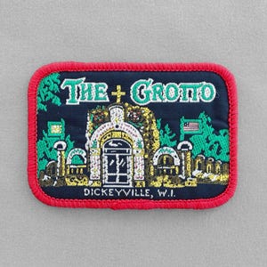 May include: A red, white, and blue embroidered patch with the text "The Grotto" and the location "Dickeyville, WI". The patch features a building with a white facade and a green roof, surrounded by green trees.