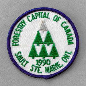 May include: Circular patch with a white center, featuring a green tree design and text. The text reads "FORESTRY CAPITAL OF CANADA" and "SAULT STE. MARIE ONT. 1990". The border is dark blue.