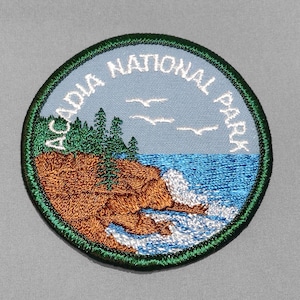 May include: A circular embroidered patch featuring a landscape of Acadia National Park. The design includes a brown rocky coastline, green pine trees, blue ocean water with white crashing waves, and white birds against a light blue sky.