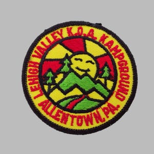 May include: Embroidered patch with a yellow background, red accents, and black border. The patch features a smiling sun, green mountains, and the text "LEHIGH VALLEY K.O.A. KAMPGROUND ALLENTOWN PA."
