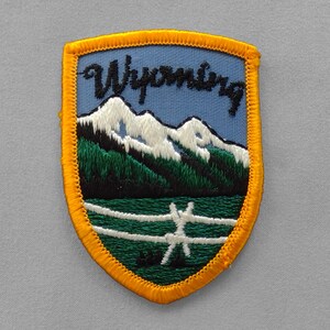May include: A shield-shaped embroidered patch featuring a light blue sky with the word Wyoming in black script. Below the text are white snow-capped mountains, a dark green forest, and a white fence set against a dark green field, all bordered by a thick yellow stitched edge.
