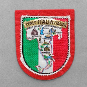 May include: A shield-shaped patch with the Italian flag colors: green, white, and red. The patch features an embroidered map of Italy with city names and the text "ITALY ITALIA ITALIEN". The patch has a red border.