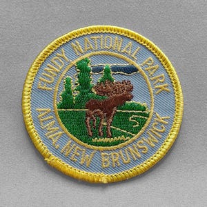 May include: A round patch with a light blue background and gold trim. The patch features the text "FUNDY NATIONAL PARK ALMA, NEW BRUNSWICK" in a circular pattern. The center shows a moose and trees.