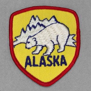 May include: A yellow felt patch with a red border in the shape of a shield. The patch features a white polar bear walking in front of a white mountain range. The text "ALASKA" is embroidered in blue below the bear.