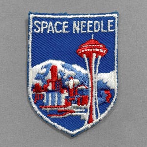 May include: Embroidered patch featuring the Space Needle in Seattle. The patch has a blue background with a white border. The Space Needle is depicted in red and white, with the text "SPACE NEEDLE" in white.