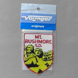 May include: A rectangular patch featuring the Mount Rushmore monument with the text "MT. RUSHMORE S.D." in red. The patch is yellow, black, and white, with a red border. The patch is in a clear plastic sleeve with a blue and white Voyager Originals header.