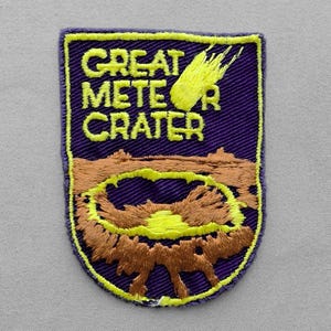 May include: A shield-shaped embroidered patch with a purple background. It features the yellow text GREAT METEOR CRATER and a yellow comet graphic above a brown crater illustration with a bright yellow center, all outlined in yellow thread.