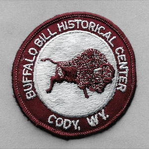May include: A circular embroidered patch featuring a dark red border with the white text BUFFALO BILL HISTORICAL CENTER and CODY, WY. The center of the patch is white and displays a dark red stitched illustration of a buffalo in profile.
