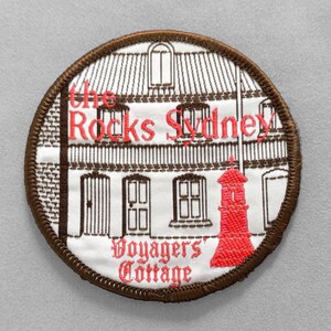 May include: Embroidered patch featuring a detailed illustration of a building with the text "The Rocks Sydney" and "Voyagers' Cottage" in red. The patch has a brown border and a white background.