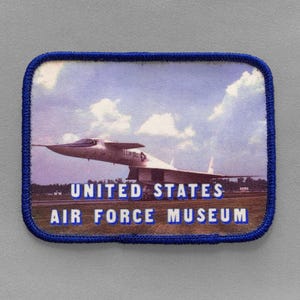 May include: A rectangular patch with a blue border features an image of a jet aircraft on a runway under a cloudy sky. The text "UNITED STATES AIR FORCE MUSEUM" is printed in white.