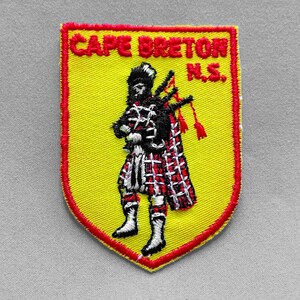 May include: Embroidered patch with a yellow background and a red border. The patch features a Scottish bagpiper in traditional attire, with the text "CAPE BRETON N.S." in red. The bagpiper is wearing a black hat and a red and white plaid kilt.