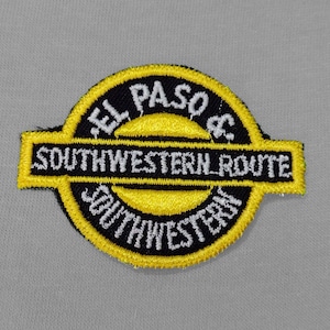 May include: Embroidered patch with the text "EL PASO & SOUTHWESTERN ROUTE" in white on a black and yellow circular design. The patch is on a light gray fabric background.