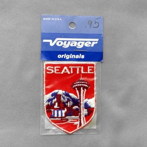 May include: A shield-shaped embroidered patch featuring a red background with the white text SEATTLE above a depiction of the Space Needle and mountain range. The patch is inside a clear plastic sleeve with a blue header card labeled Voyager originals.
