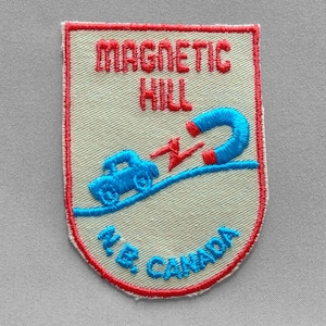 May include: Embroidered patch with a red border and the words "MAGNETIC HILL" in red. A blue car is depicted on a blue road, with a blue and red magnet above it. The text "N.B. CANADA" is at the bottom.