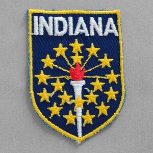 May include: A blue and yellow embroidered patch with the word "INDIANA" in white lettering. The patch features a torch with a red flame and 19 yellow stars.