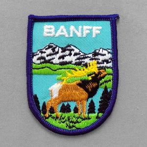 May include: Embroidered patch featuring the word "BANFF" above a mountain scene with a lake, trees, and a moose. The patch has a blue border and a shield shape. The moose is brown and black with yellow antlers.