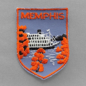 May include: Embroidered patch featuring the word "MEMPHIS" above a riverboat scene. The patch has a light blue background with orange and black accents. The patch is shield-shaped.