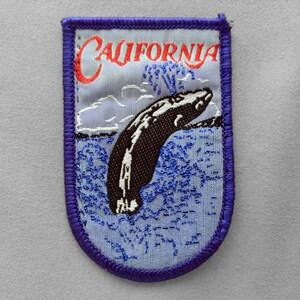 May include: A rectangular embroidered patch with a purple stitched border. The design features a light blue background with a dark brown whale breaching the water, white clouds, and the red text California arched across the top section.