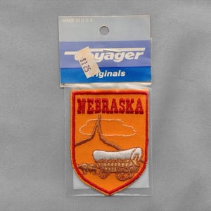 May include: A shield-shaped embroidered patch featuring an orange background with a red border. The text NEBRASKA is stitched in red at the top. Below, a brown covered wagon with white canvas is depicted. The patch is in a clear plastic bag.