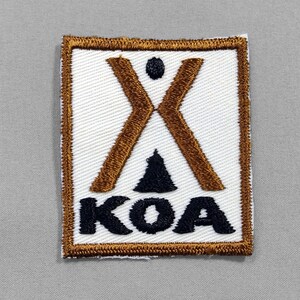 May include: Square embroidered patch with a white background, featuring a brown 'X' shape, a black dot, a black triangle, and the letters 'KOA' in black. The patch has a brown border.