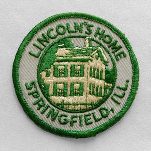 May include: Embroidered patch featuring a detailed illustration of a house, likely Lincoln's Home, in Springfield, IL. The design is in shades of green and tan, with the text "LINCOLN'S HOME SPRINGFIELD, ILL." in a circular arrangement around the house.
