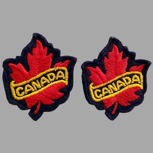 May include: Two embroidered patches shaped like red maple leaves with the word "CANADA" in yellow lettering. The leaves have a dark blue border and are set against a gray background. These patches are likely for clothing or accessories.