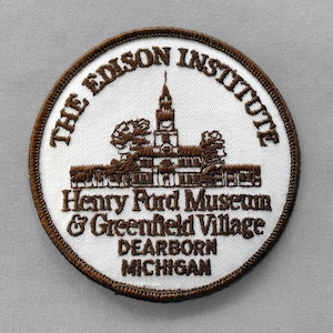 May include: A circular patch with a brown border and white center. The text "THE EDISON INSTITUTE" arches across the top. Below is a brown illustration of a building and the text "Henry Ford Museum & Greenfield Village DEARBORN MICHIGAN".