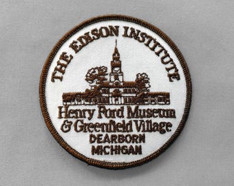 May include: A circular patch with a brown border and white center. The text "THE EDISON INSTITUTE" arches across the top. Below is a brown illustration of a building and the text "Henry Ford Museum & Greenfield Village DEARBORN MICHIGAN".