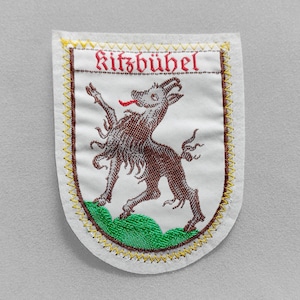 May include: Embroidered patch featuring a brown goat-like creature with a red tongue, standing on green grass. The word "Kitzbühel" is embroidered in red above the creature. The patch has a white background with a yellow and brown stitched border.