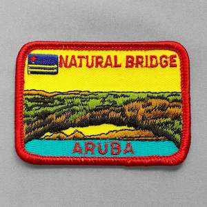 May include: Embroidered patch featuring a scenic view of Aruba's Natural Bridge. The patch has a yellow background with the words "NATURAL BRIDGE" and "ARUBA" in red. The design includes a blue and yellow flag, green and brown landscape, and blue water, all framed by a red border.