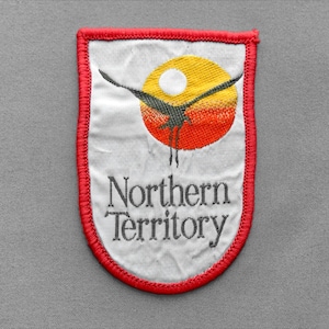May include: A shield-shaped embroidered patch with a white background and a red stitched border. It features a black bird silhouette flying over a yellow and orange sun. The text Northern Territory is embroidered in grey below the graphic.