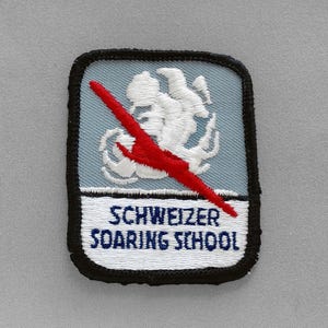 May include: Embroidered patch with a light blue background, a white graphic, and a red design. The patch has the words "SCHWEIZER SOARING SCHOOL" in white text on a white background, framed by a black border.