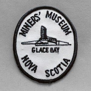 May include: A circular patch with a black border and white center. The patch features the text "MINERS' MUSEUM" and "GLACE BAY NOVA SCOTIA" in black. A black and white graphic of a building is in the center.