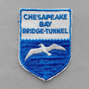May include: Embroidered patch featuring the text "CHESAPEAKE BAY BRIDGE-TUNNEL" in white on a blue and white shield design. A white seagull flies over blue waves. The patch has a white border.