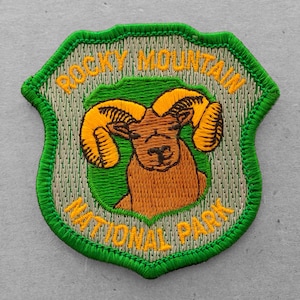 May include: Green and beige embroidered patch with the words "Rocky Mountain National Park" and a brown and yellow bighorn sheep.