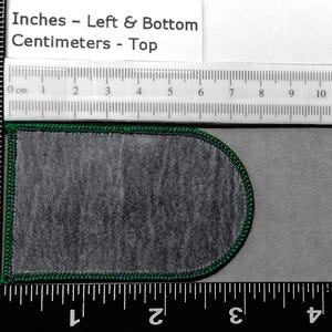 May include: A green-trimmed, gray fabric patch with a rounded top and bottom. The patch is measured in inches and centimeters using a ruler.