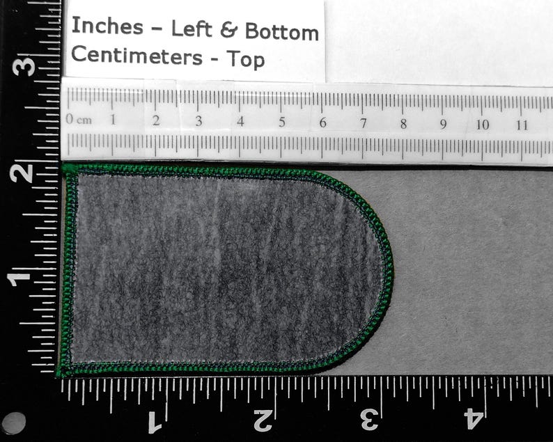 May include: A green-trimmed, gray fabric patch with a rounded top and bottom. The patch is measured in inches and centimeters using a ruler.