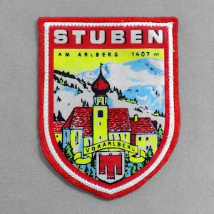 May include: Embroidered patch with a red border and a shield shape. The patch features the text "STUBEN AM ARLBERG 1407 m" above a colorful illustration of a church and snowy mountains. The text "VORARLBERG" is on a yellow banner.