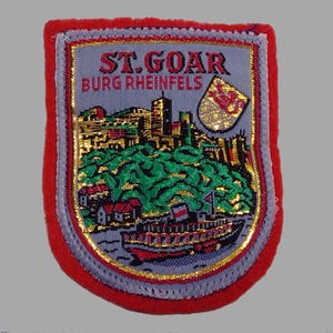 May include: Embroidered patch with a red border, featuring the text "ST. GOAR BURG RHEINFELS". The patch depicts a landscape with a riverboat, buildings, and a shield emblem. The design incorporates gold, green, and red colors.