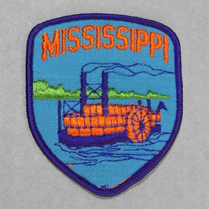 May include: A blue and purple embroidered patch with the word "MISSISSIPPI" in orange lettering. The patch features a steamboat with orange and blue details on a blue river.
